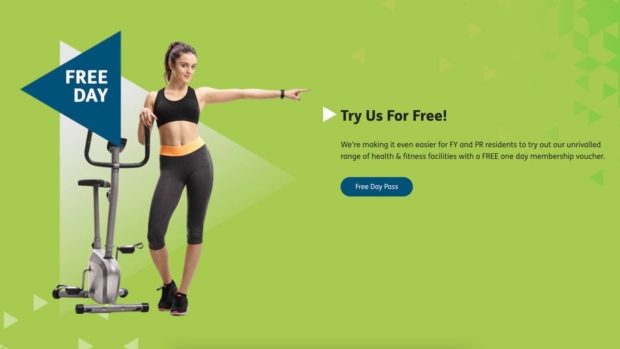 Woman in gym wear standing next to an exercise bike, pointing toward a 'Free Day' banner, with heading 'Try Us For Free!' promoting a free one-day membership voucher for FY and PR residents at YMCA Fylde Coast and a 'Free Day Pass' button.
