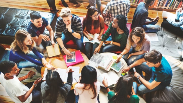 Aerial view of a diverse group of students sitting around a table in a library, discussing and studying together with books, laptops, and notes.